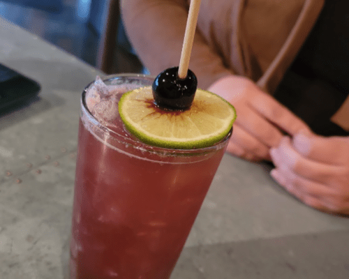 A close-up of a cocktail with a lime slice and cherry on top, served in a glass with ice. A hand is visible in the background.