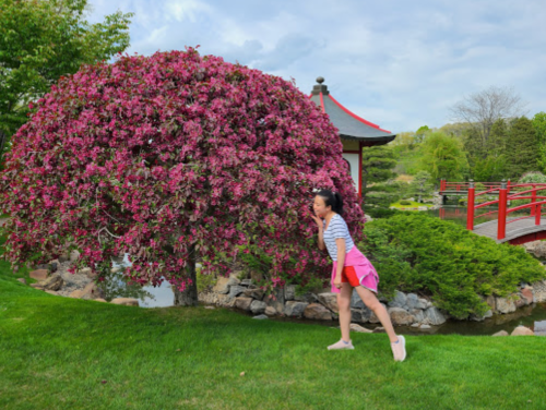 A woman in a pink skirt playfully leans towards a vibrant pink flowering tree in a serene garden setting.
