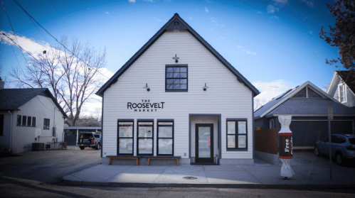 A white, modern building with a sloped roof, featuring "The Roosevelt Market" sign and large windows.