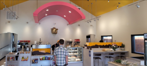 A bright café interior with a pink arch, a display case, and a customer at the counter. Decor includes a cow head.