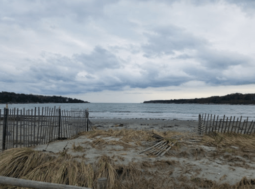 A cloudy beach scene with a view of the water, framed by wooden fencing and patches of dry grass.