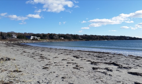 A sandy beach with gentle waves, surrounded by trees and houses under a clear blue sky with scattered clouds.