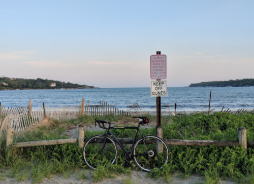 A bicycle parked near a beach with a "Keep Off Dunes" sign and a view of the water and distant shoreline.