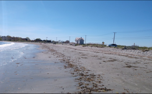 A sandy beach with gentle waves, a house in the distance, and power lines under a clear blue sky.