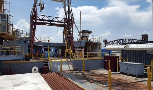 A view of an industrial ship deck with equipment, railings, and a bridge in the background under a blue sky.