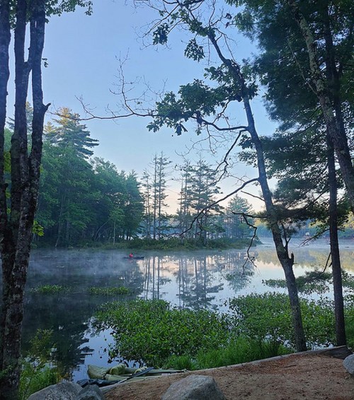A serene lake surrounded by trees, with mist rising from the water and a kayaker in the distance.