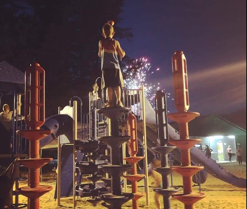 A child stands on a playground structure at night, with fireworks lighting up the sky in the background.