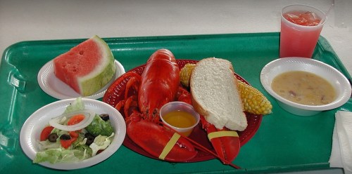 A vibrant seafood meal featuring a lobster, corn, salad, bread, watermelon, and a drink on a green tray.