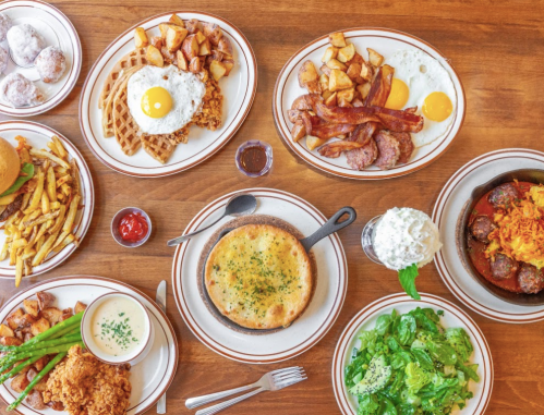 A spread of various dishes including fried chicken, waffles, eggs, bacon, salad, and desserts on a wooden table.