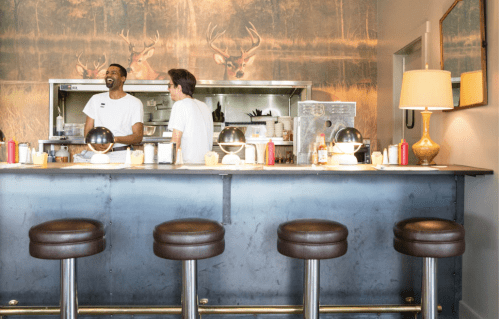 Two people at a diner counter with a deer-themed mural in the background and retro-style bar stools.