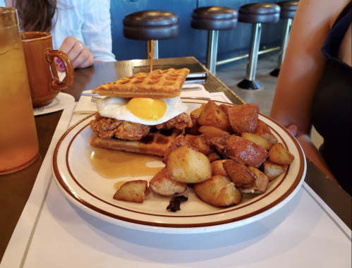 A plate with a chicken and waffle sandwich topped with a fried egg, served with roasted potatoes and a drink.