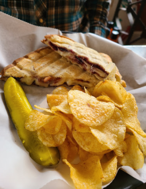 A grilled sandwich with layers of meat and cheese, served with potato chips and a pickle on the side.