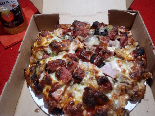 A pizza with various toppings, including meats and vegetables, in a cardboard box on a red table.