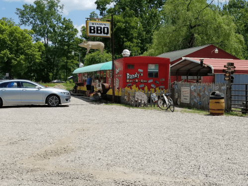 A colorful BBQ restaurant with a cow sign, surrounded by gravel parking and greenery. A car and bicycle are visible.