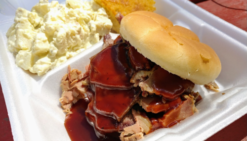 A plate with a pulled pork sandwich topped with barbecue sauce, a side of potato salad, and a cornbread muffin.