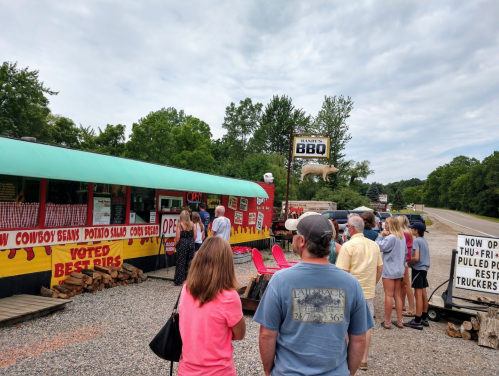 A line of people waiting outside a BBQ restaurant with a sign, surrounded by trees and a gravel parking area.