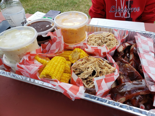 A tray of barbecue featuring pulled chicken, ribs, corn, and three sauces in cups, served on a picnic table.