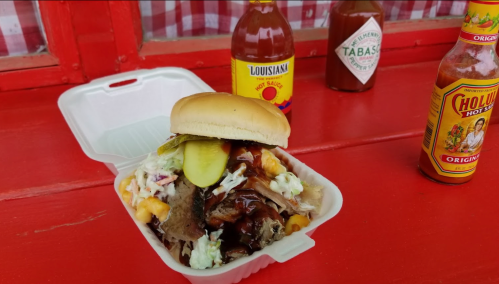 A pulled pork sandwich with coleslaw and pickles, served in a takeout container, alongside hot sauce bottles.