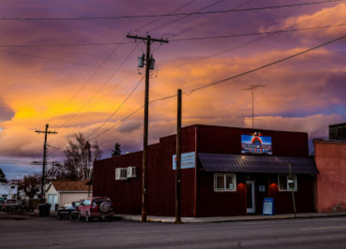 A red building with a sign under a colorful sunset, surrounded by power lines and a few trees.