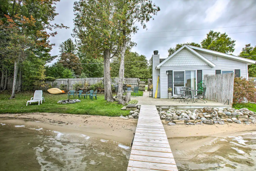A cozy lakeside cottage with a wooden dock, surrounded by trees and outdoor seating on a sandy shore.