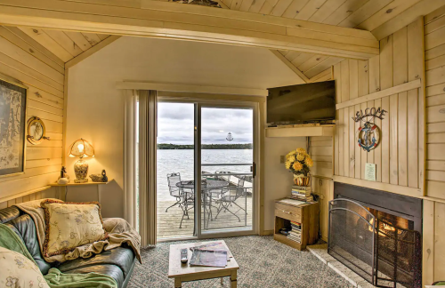 Cozy cabin interior with a sofa, fireplace, and large windows overlooking a lake and outdoor seating area.