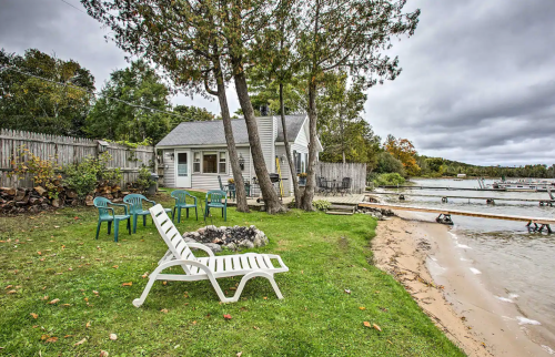 A cozy cottage by the water, surrounded by chairs and trees, with a sandy beach and cloudy sky.