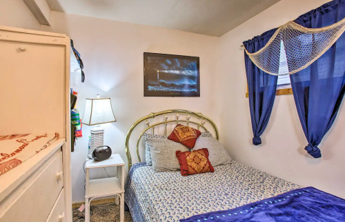 Cozy bedroom with a bed, decorative pillows, a lamp, and blue curtains, featuring a lighthouse painting on the wall.