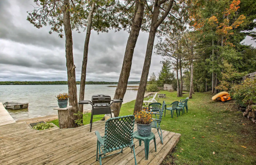 A serene lakeside view with a wooden deck, chairs, a grill, and trees, set against a cloudy sky.