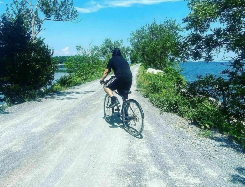 A person rides a bicycle along a gravel path by a lake, surrounded by trees and blue skies.