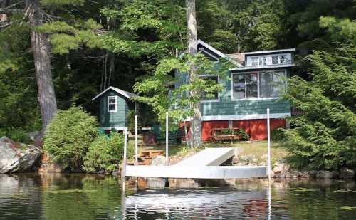 A green cottage by a lake, surrounded by trees, with a small dock and a picnic area visible in the foreground.