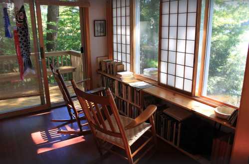 A cozy room with two rocking chairs, a bookshelf, and large windows overlooking greenery. Soft light filters in.