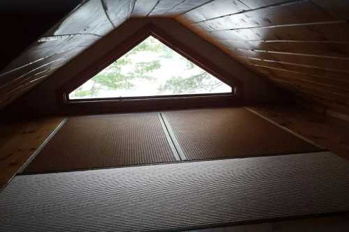 View from an attic with a triangular window overlooking trees, featuring wooden walls and tatami-style flooring.