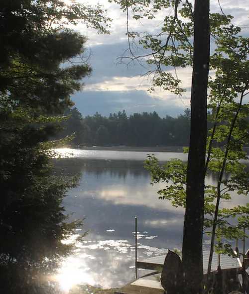 A serene lake scene with trees, a cloudy sky, and a wooden dock reflecting sunlight on the water's surface.