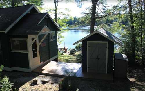 A cozy green cabin and shed by a serene lake, surrounded by trees and sunlight.