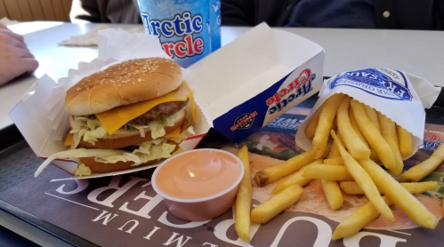 A tray with a cheeseburger, fries, and a dipping sauce, alongside a large drink in a fast-food setting.