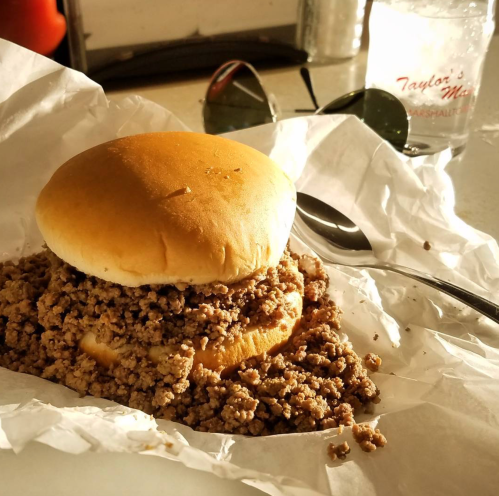 A sandwich with a soft bun filled with seasoned ground meat, served on white paper with a drink and sunglasses in the background.