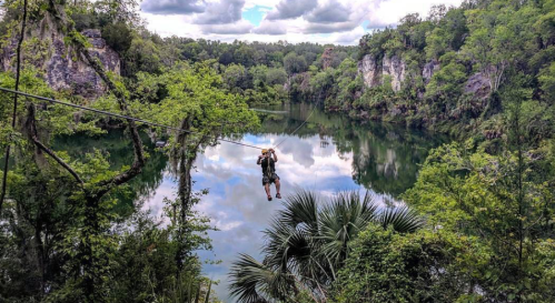 A person zip-lining over a serene lake surrounded by lush greenery and rocky cliffs under a cloudy sky.