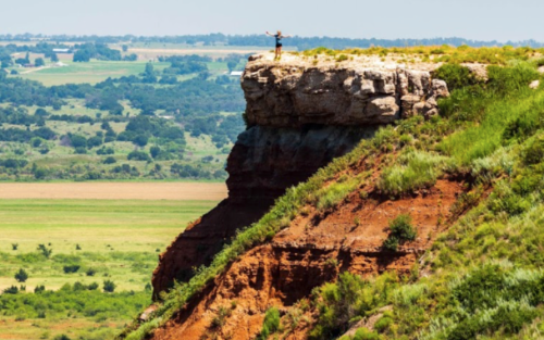 A person stands triumphantly on a rocky cliff, overlooking a vast green landscape below.