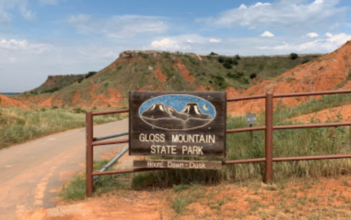 Sign for Gloss Mountain State Park at the entrance, with red rock formations and green grass in the background.