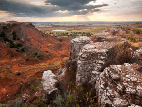 A scenic view of rugged cliffs overlooking a vast, colorful landscape under a dramatic sky with rays of sunlight.