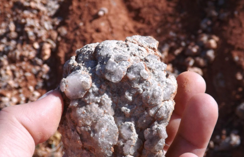 A hand holding a rough, mineral-rich rock with a textured surface against a background of soil and gravel.
