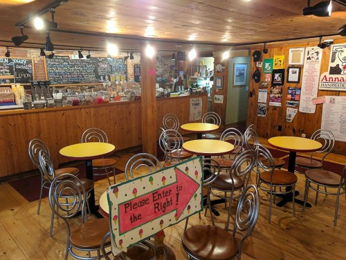 Cozy café interior with yellow tables, metal chairs, a counter with a menu, and a sign directing to the right.