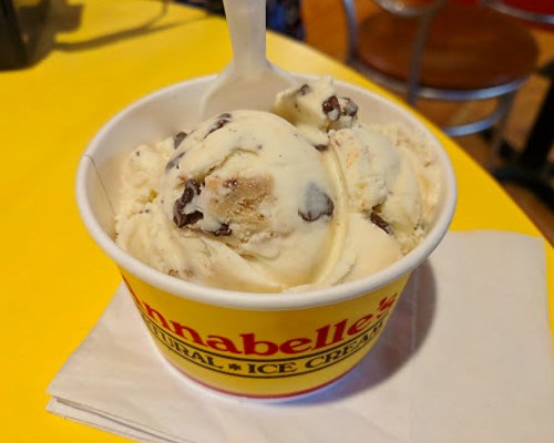 A cup of ice cream with chocolate chip cookie dough and chocolate chunks, sitting on a yellow table.