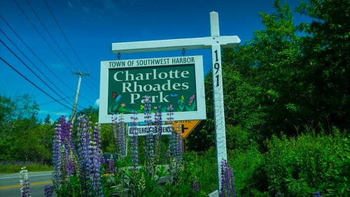 Sign for Charlotte Rhoades Park in Southwest Harbor, surrounded by greenery and purple flowers under a clear blue sky.