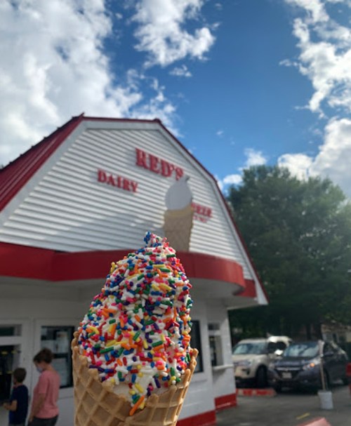 A colorful ice cream cone with sprinkles in front of a dairy shop named "Red's" under a blue sky.