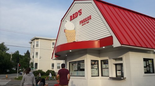 A small ice cream shop with a red roof and a sign reading "Red's Treats" as people walk towards it.