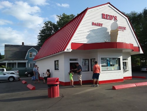A small ice cream shop with a red and white roof, featuring customers ordering at the window on a sunny day.
