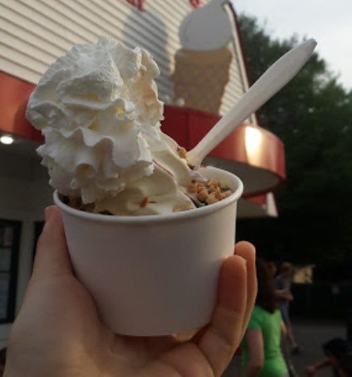 A hand holds a cup of ice cream topped with whipped cream and nuts, with a shop in the background.