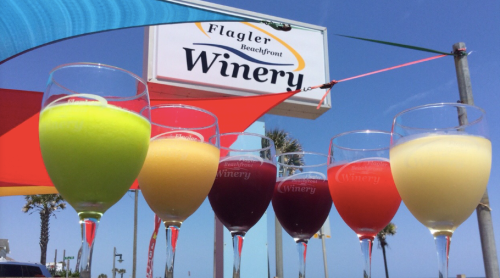 Colorful drinks in glasses in front of a winery sign, with vibrant flags and a clear blue sky in the background.