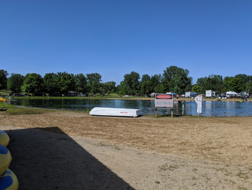 A sandy beach area by a calm lake, with trees in the background and a warning sign near the water's edge.